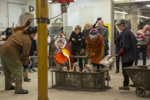 Photo of a a team of five people handling heavy equipment to pour molten bronze into molds. Behind the team stand five people looking intently at the pour in front of them. 