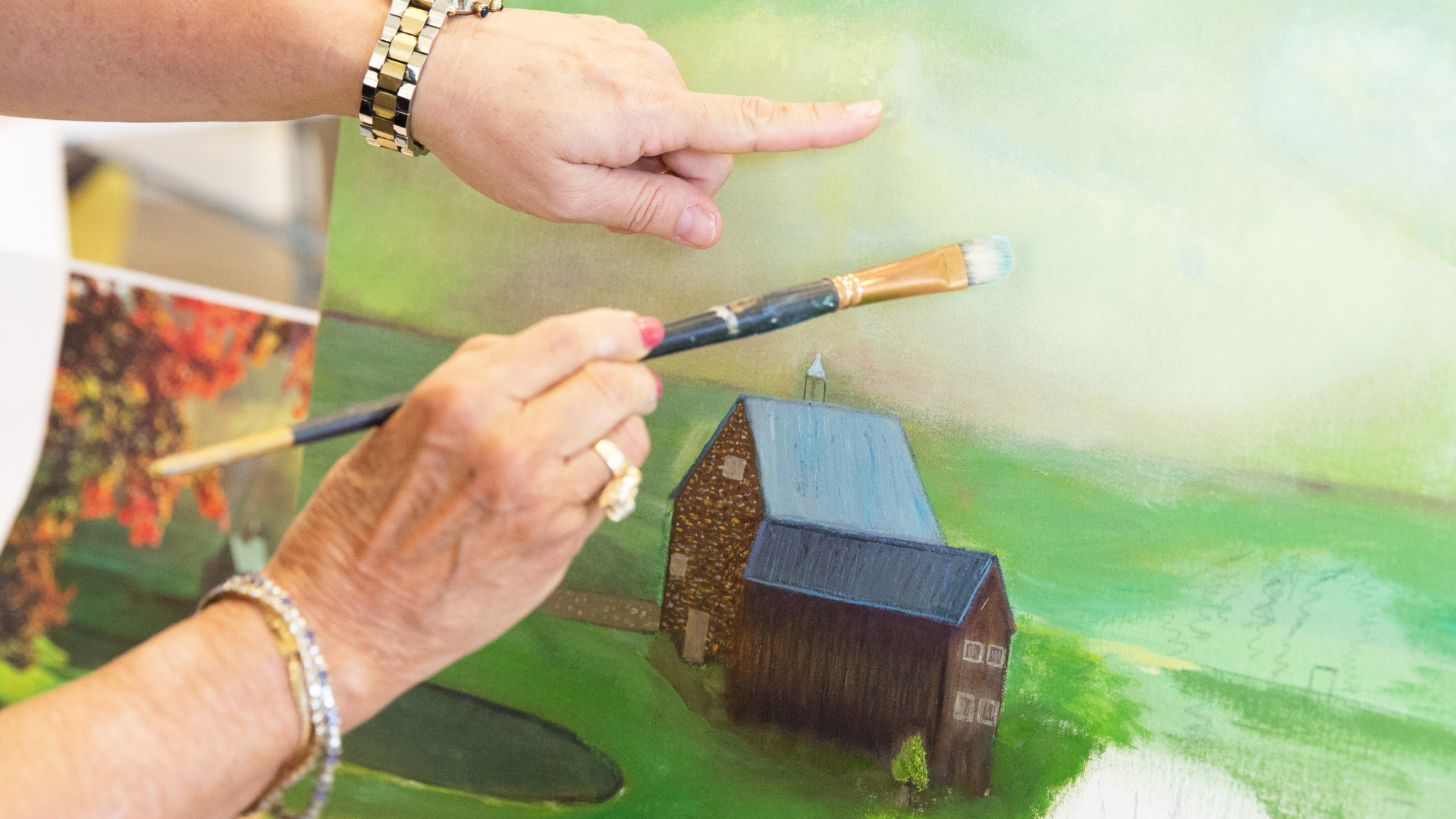 One person is holding a paintbrush and actively applying paint to the canvas, while the other is pointing, possibly providing instruction or feedback.