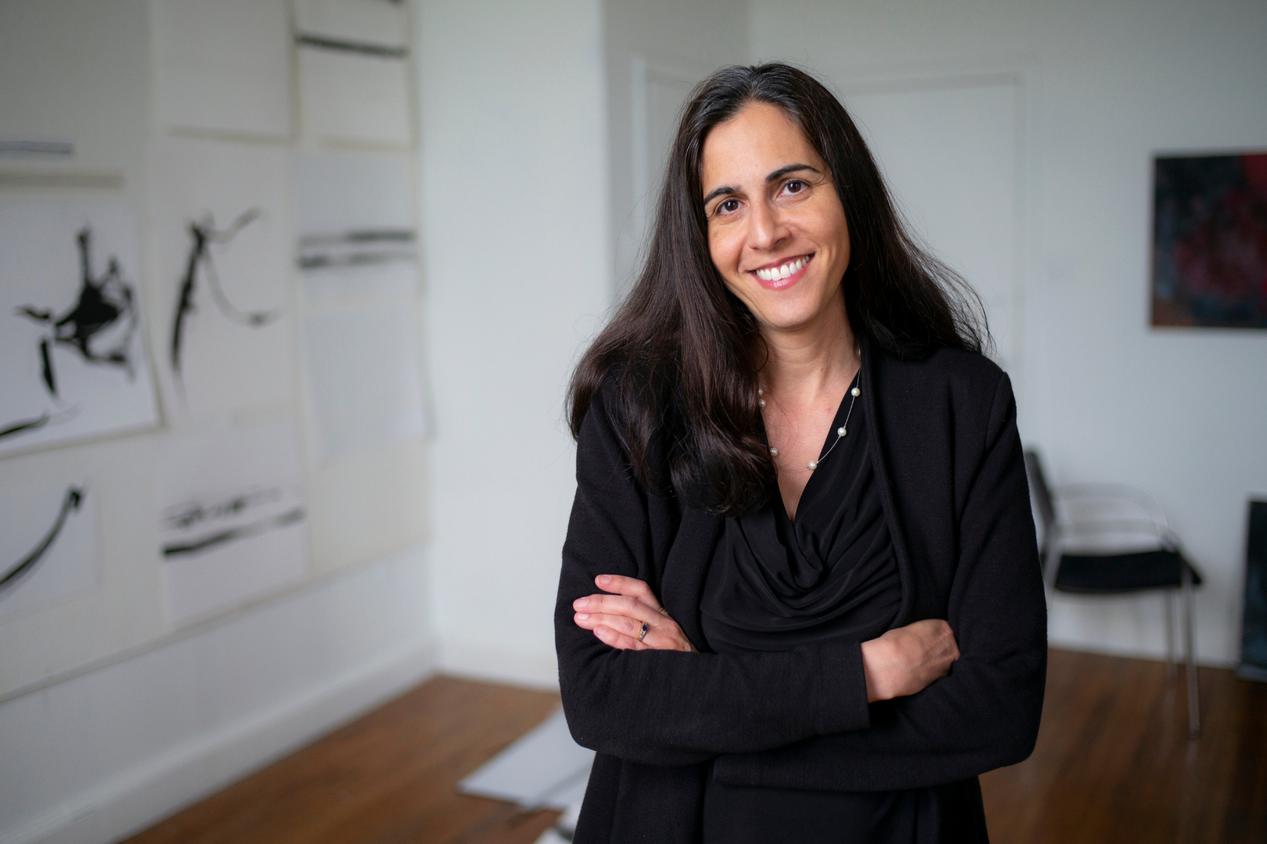 Dr. Nazanin Moghbeli, a cardiologist and artist, stands in a brightly lit studio with her arms crossed, wearing a black top and blazer in front of walls adorned with minimalist artwork.