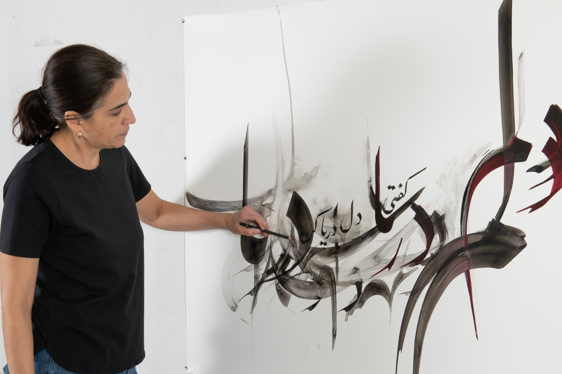 A woman stands before a large white canvas, using traditional tools to create an abstract drawing inspired by Persian calligraphy.