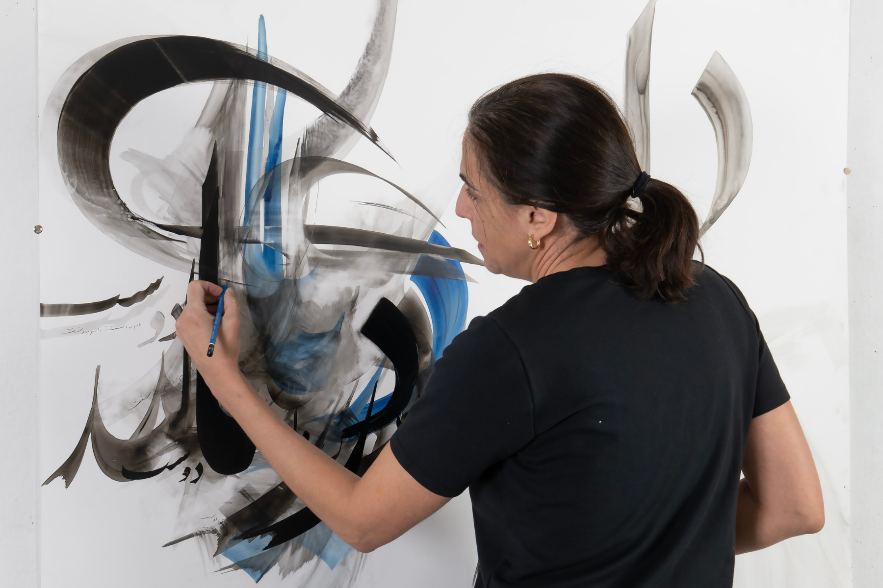 An artist with dark hair tied back in a ponytail and wearing a black t-shirt is captured from behind as she creates large-scale, expressive black and blue calligraphic strokes on a white canvas.