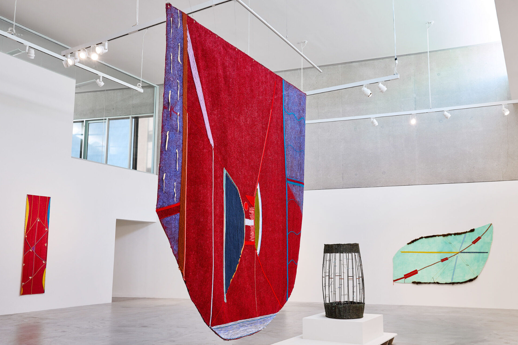 A large red, blue, and purple textile artwork hangs from the ceiling in a bright contemporary gallery, surrounded by additional colorful fabric pieces on the walls and a woven sculptural object displayed on a pedestal.