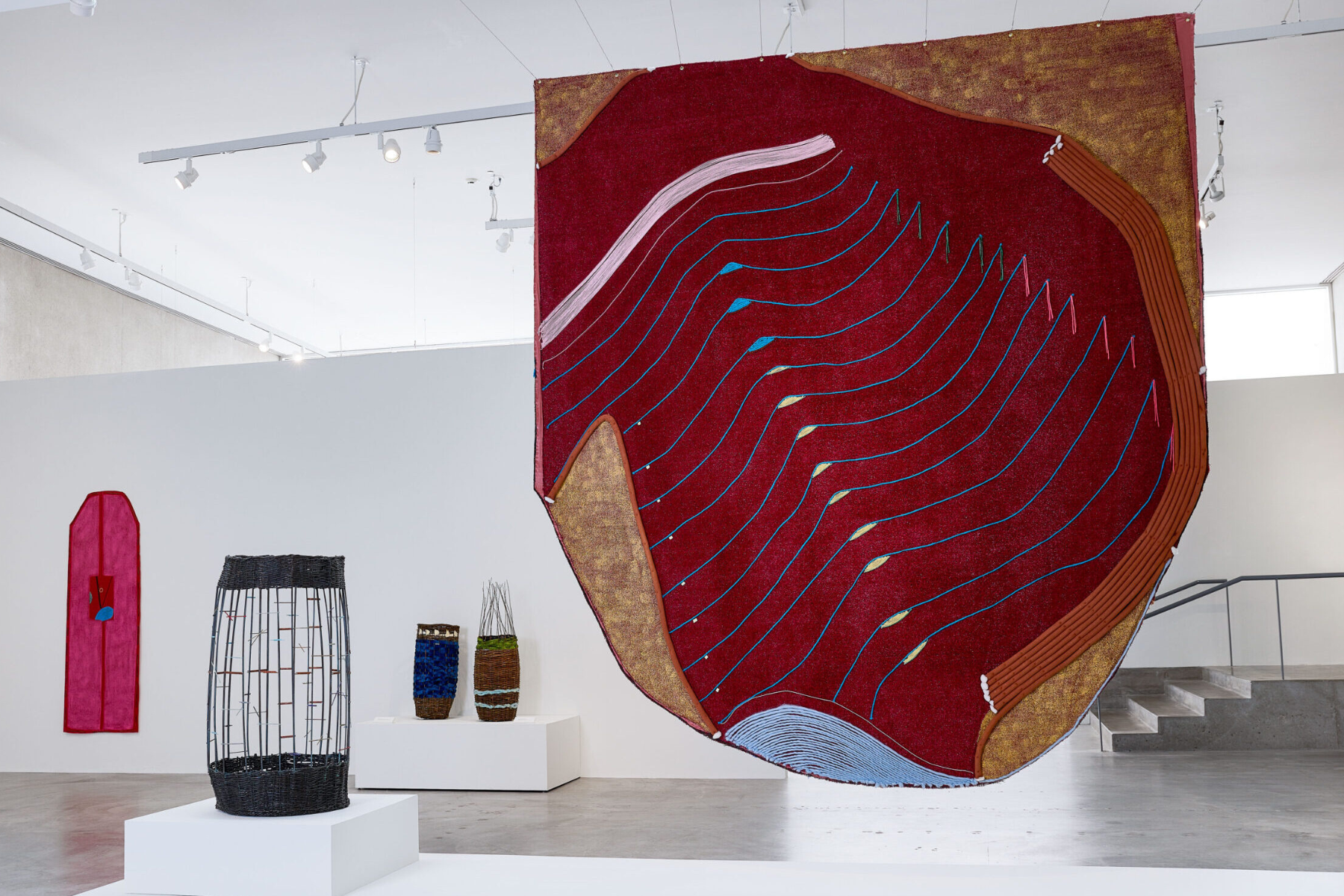 A large red and gold textile artwork with flowing blue and white stitched lines hangs prominently in a bright gallery, accompanied by additional woven vessels on pedestals and colorful fabric works on the surrounding walls.