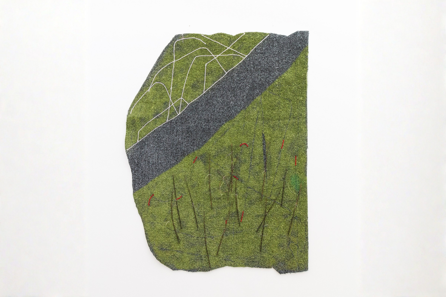 A green and gray irregularly shaped textile artwork hangs on a white wall, featuring stitched white lines in the upper section and scattered red and dark vertical marks across the lower area.