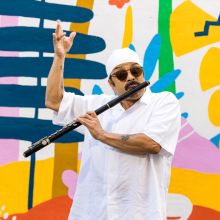 Photo of Eubie Nieves playing a black flute in front of a colorful mural. Eubie is wearing all white, with a white headscarf. He is wearing sunglasses. 